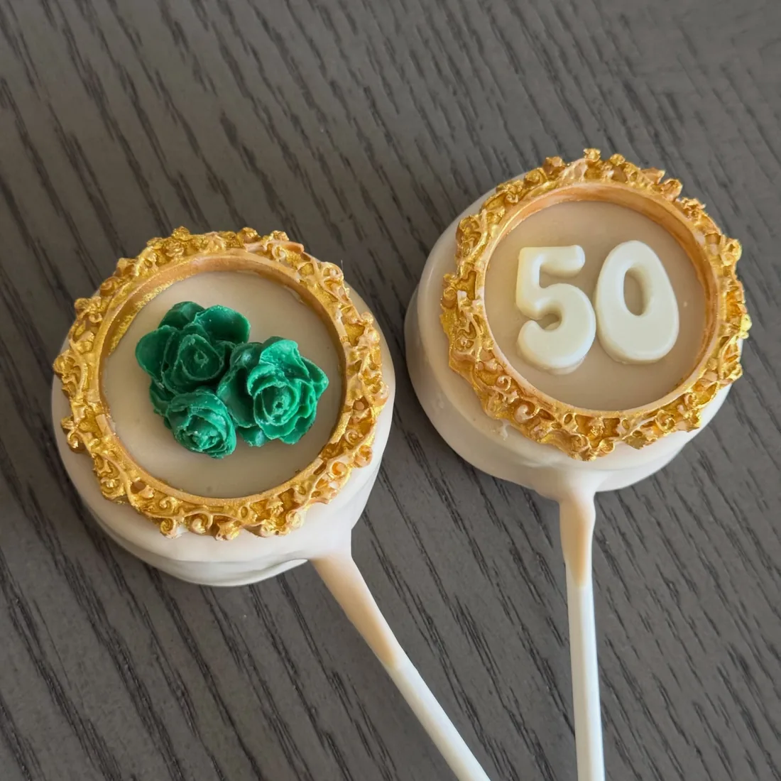 Two gold-rimmed cake pops, one with emerald roses and one with a fondant 50