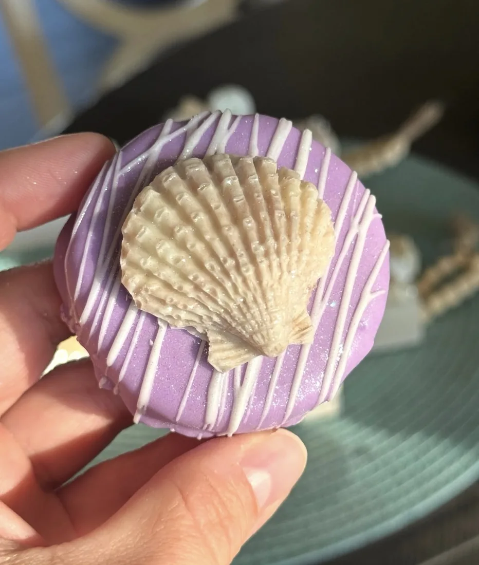 Hand holding a purple dipped cake puck topped with a large white chocolate scallop shell