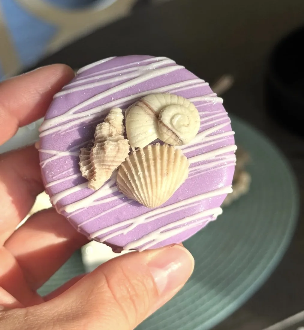 Hand holding a purple dipped cake puck topped with a cluster of white chocolate seashells