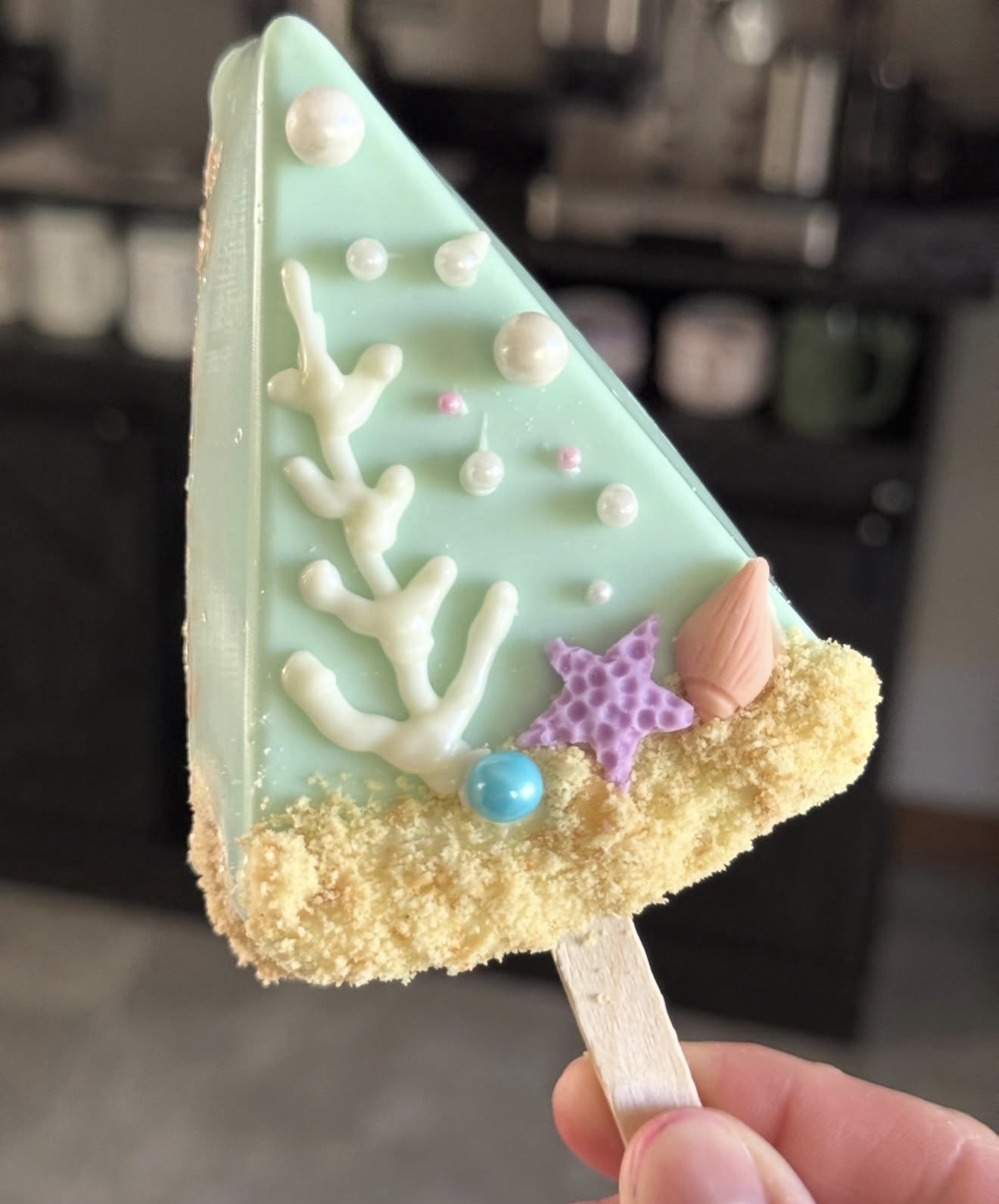 Mint rice crispy treat dipped in white chocolate with coral, purple starfish, and shell on sandy crumble base