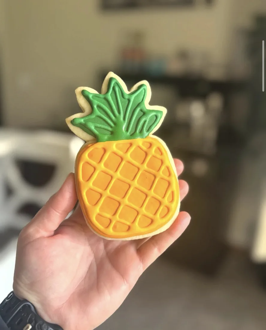Hand holding a pineapple shaped cookie decorated in yellow and green royal icing