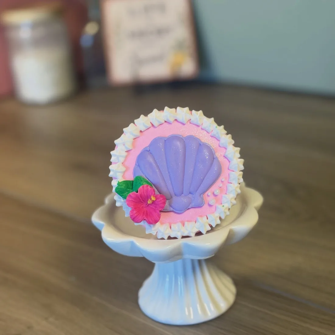 Round cookie flooded in pink royal icing with a piped shell border, a purple fondant scallop shell, a pink hibiscus and sugar pearls