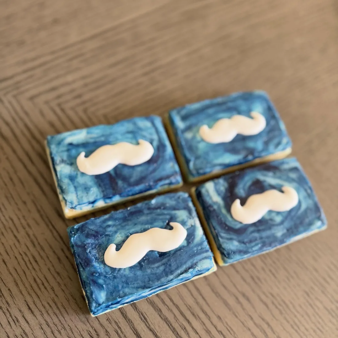 Four square cookies in blue watercolor icing with white fondant mustaches